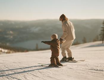 Parent and toddler on a groomed run above a lake, the child with arms out for balance