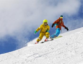 An adult and child snowboard down a mountain on a clear day