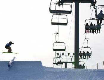 A skiier jumps from a terrain park obstacle while a ski lift operates overhead 