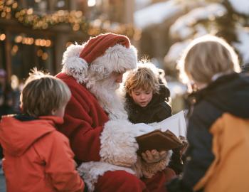Santa reading a story to children outside a holiday-lit mountain village