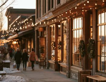 Shoppers passing glowing storefronts strung with holiday lights on a snowy mountain town sidewalk