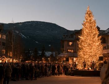 Crowd gathered around a lit Christmas tree and live band in a mountain town at dusk