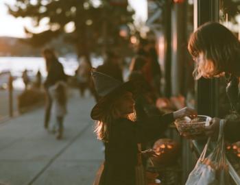 Girl in a witch hat taking candy from a shopkeeper on a lakeside sidewalk at golden hour