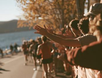 Spectators reaching out to high-five runners on a lakeside road lined with golden fall trees
