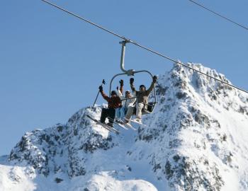 Friends cheering on a chairlift beneath a snowy granite peak and clear blue sky