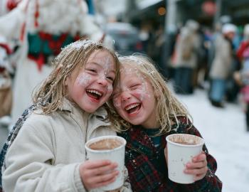 Two laughing kids with snowflake face paint holding hot cocoa at a snowy winter festival
