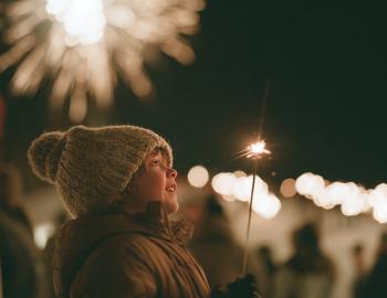 Child in a knit hat holding a sparkler and watching New Year's Eve fireworks