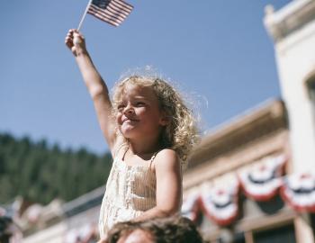 Girl on a parent's shoulders waving an American flag on a historic main street hung with bunting