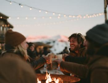 Friends laughing and toasting mugs around a fire pit under string lights in a ski village at dusk