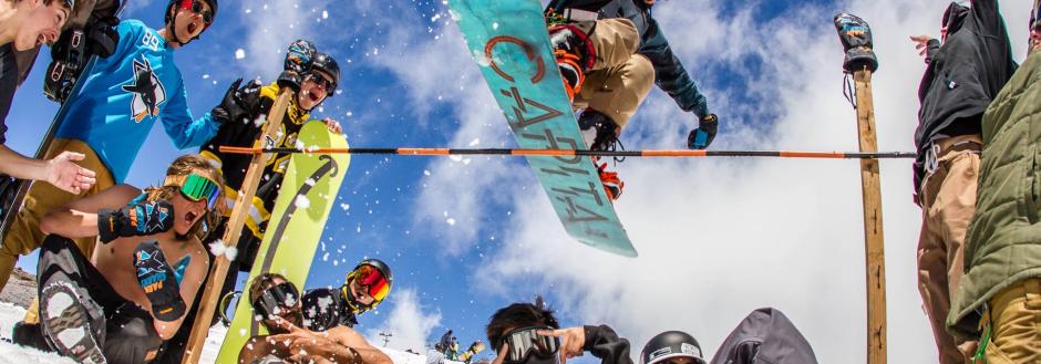 snowboarder jumping over a high stick, people encouraging snowboarder to take a high jump