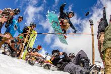 snowboarder jumping over a high stick, people encouraging snowboarder to take a high jump